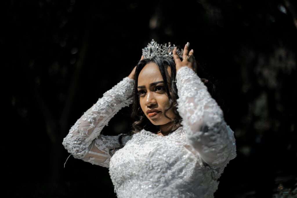 A bride in a white lace gown adjusts her tiara outdoors during a sunny daytime wedding ceremony.