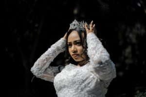 A bride in a white lace gown adjusts her tiara outdoors during a sunny daytime wedding ceremony.