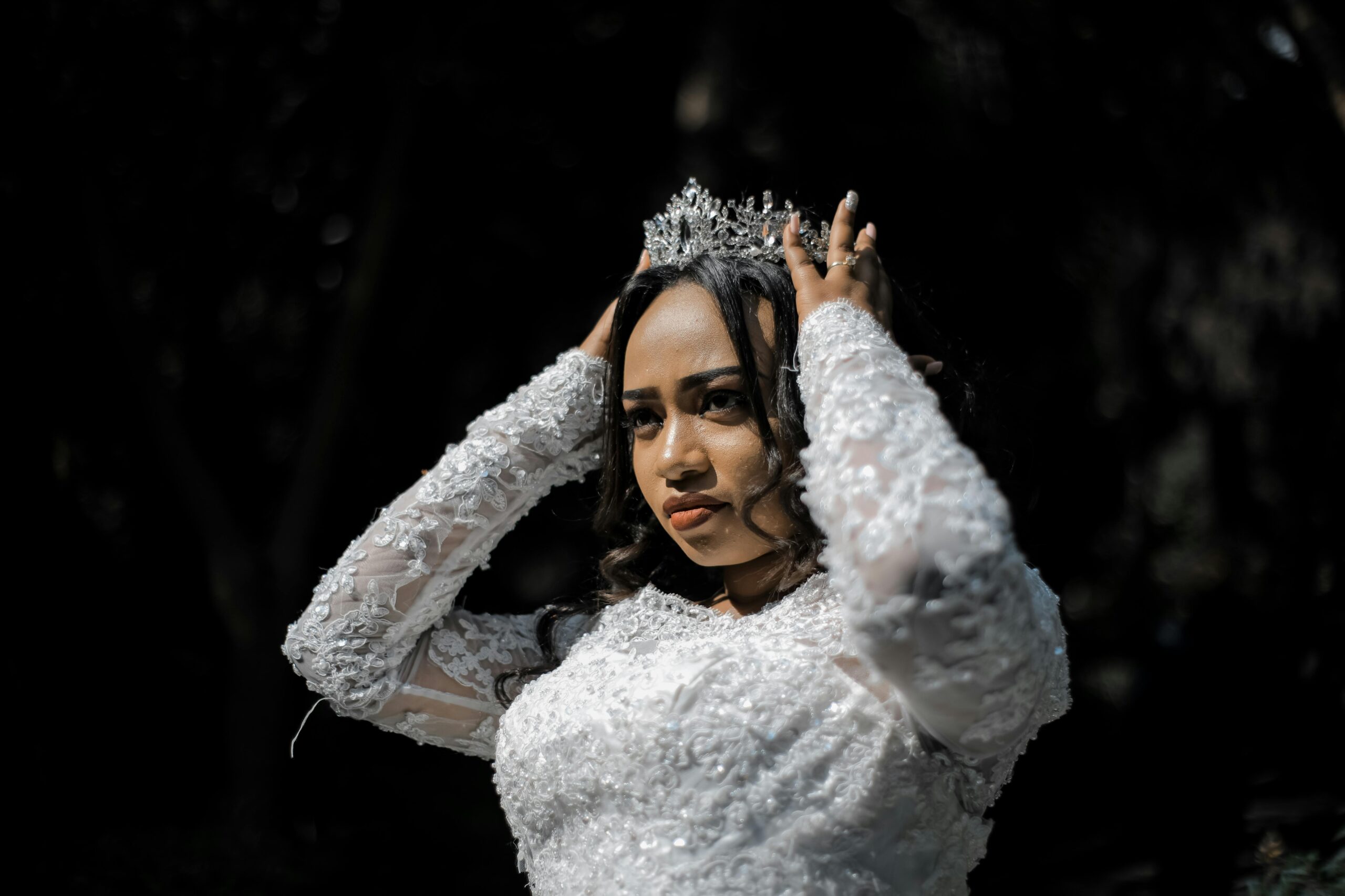 A bride in a white lace gown adjusts her tiara outdoors during a sunny daytime wedding ceremony.